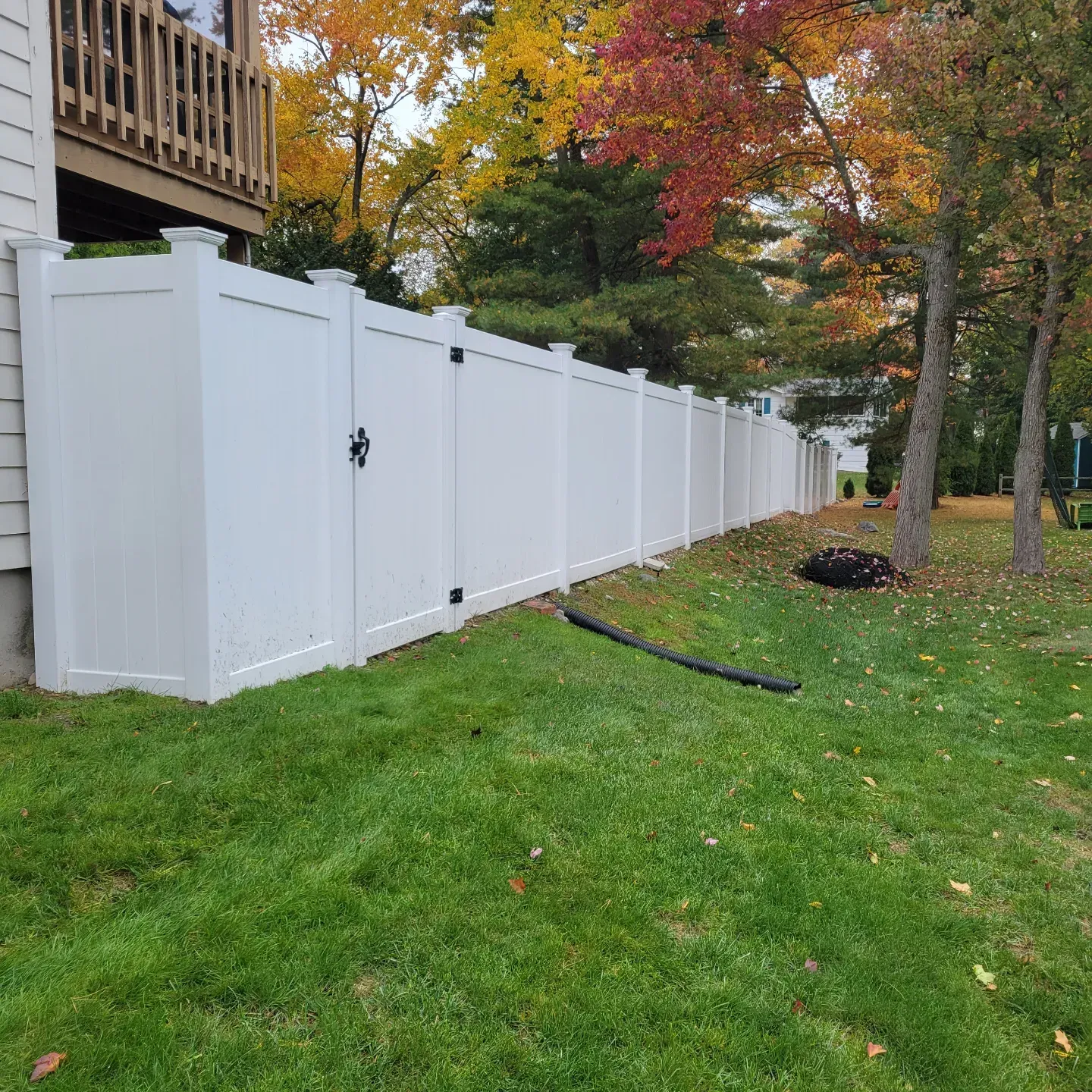 White vinyl fence bordering a grassy lawn with a gate. Fall foliage in the background.