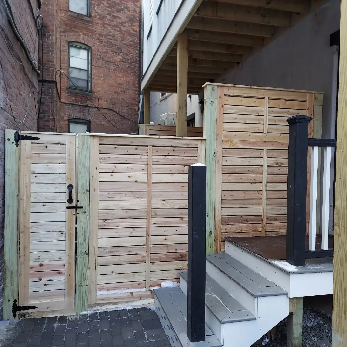 Wooden gate and fence with steps leading to a deck, between brick and light-colored walls.