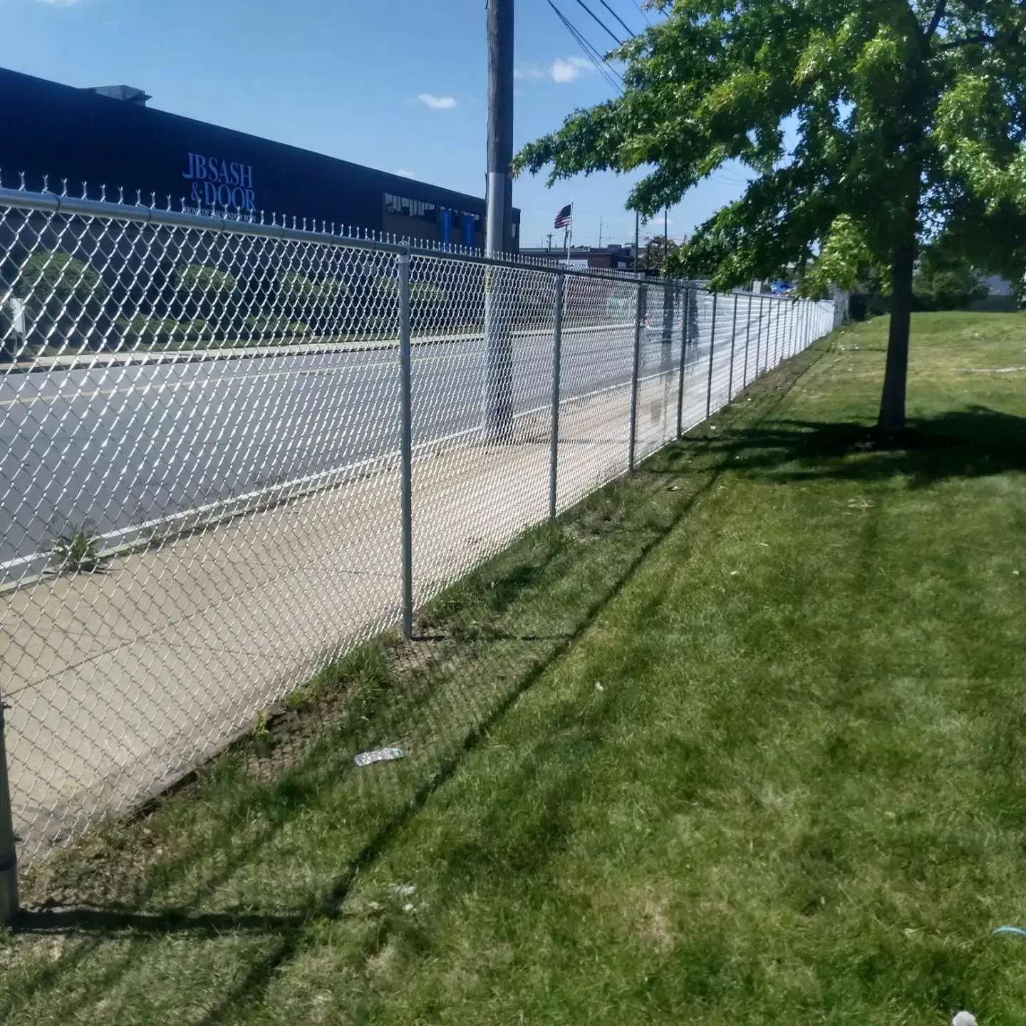 Chain-link fence along sidewalk and grass. A building and tree are visible in the background.