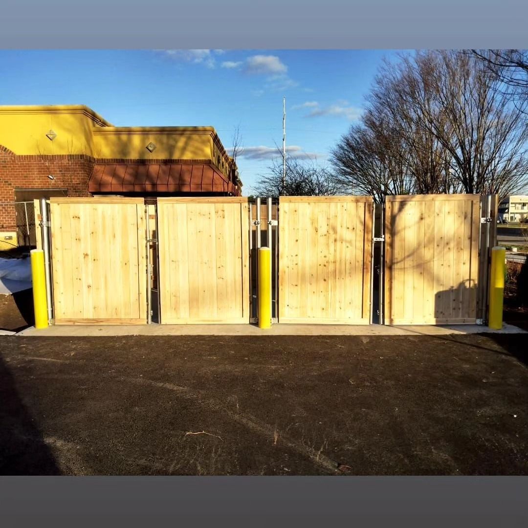 Wooden dumpster enclosure with yellow poles, in front of a building with blue sky.