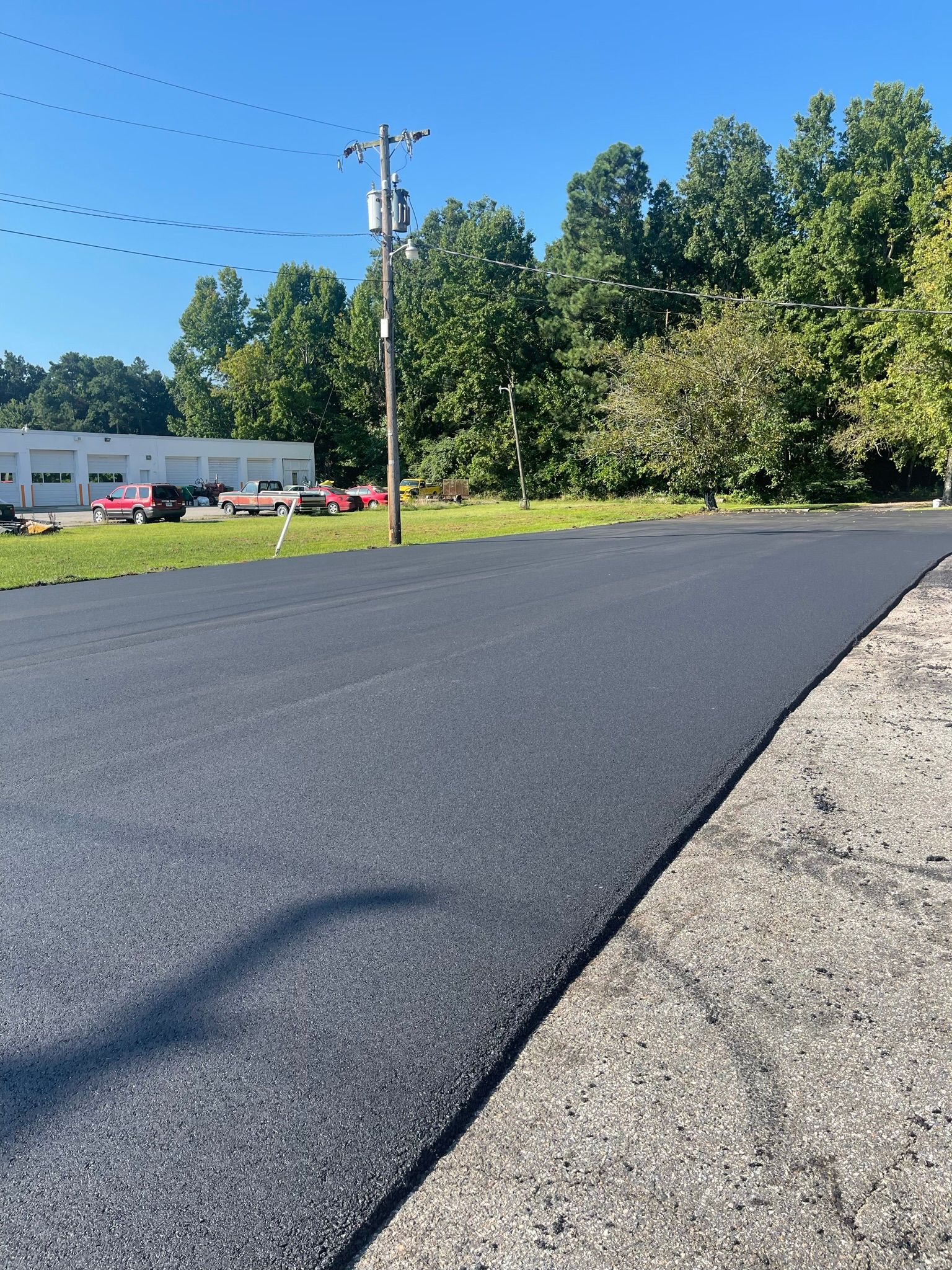 Freshly paved dark asphalt road beside a gravel shoulder, with trees, utility poles, and buildings in the background