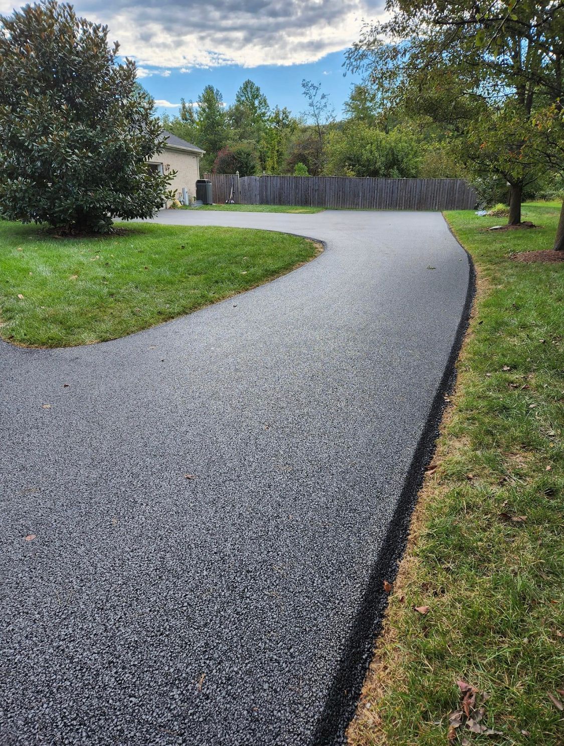 Curving gravel driveway through a green yard toward a wooden fence and trees under a blue sky
