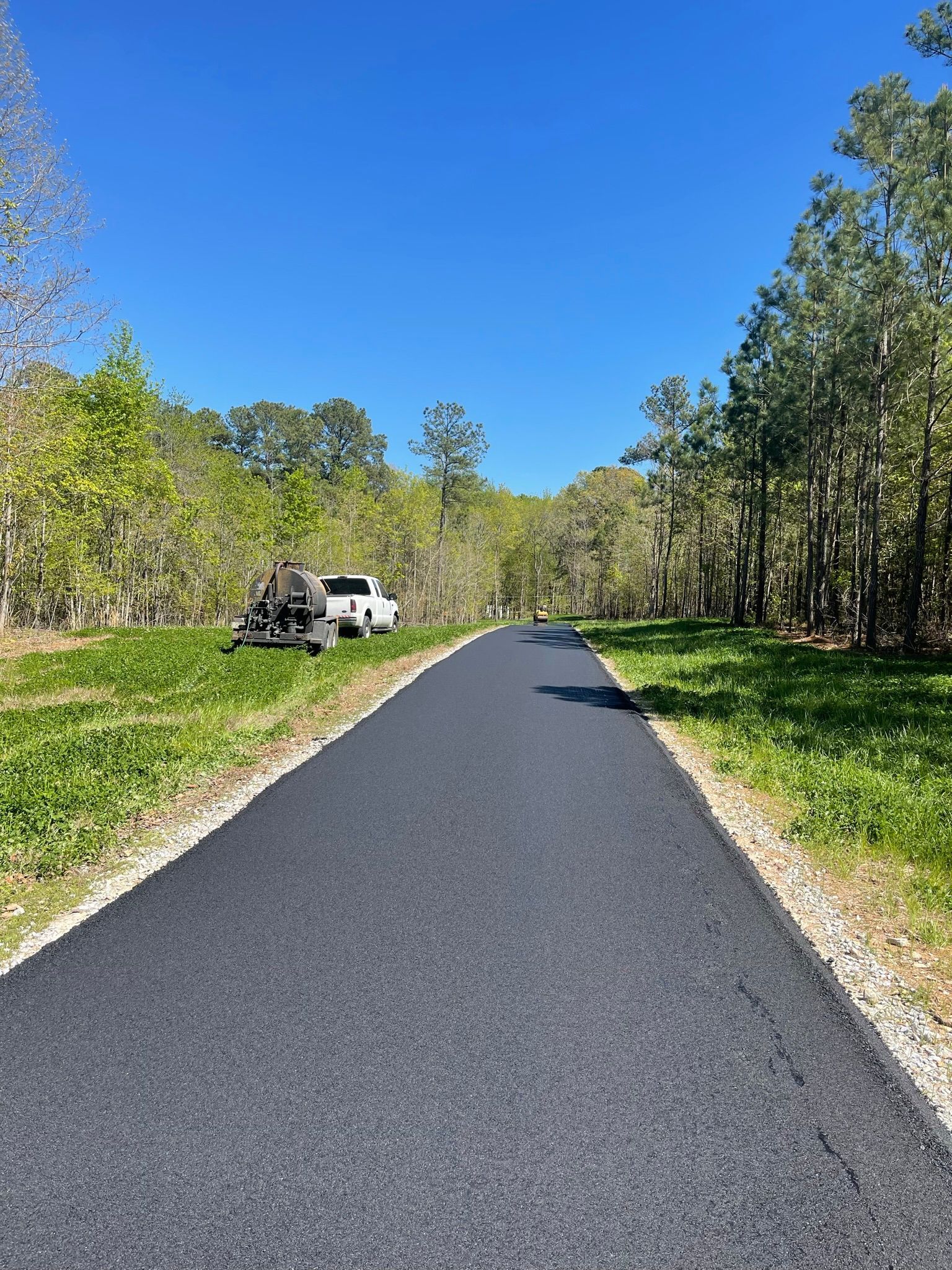 Paved road through a wooded area under a clear blue sky, with a white vehicle parked on the left