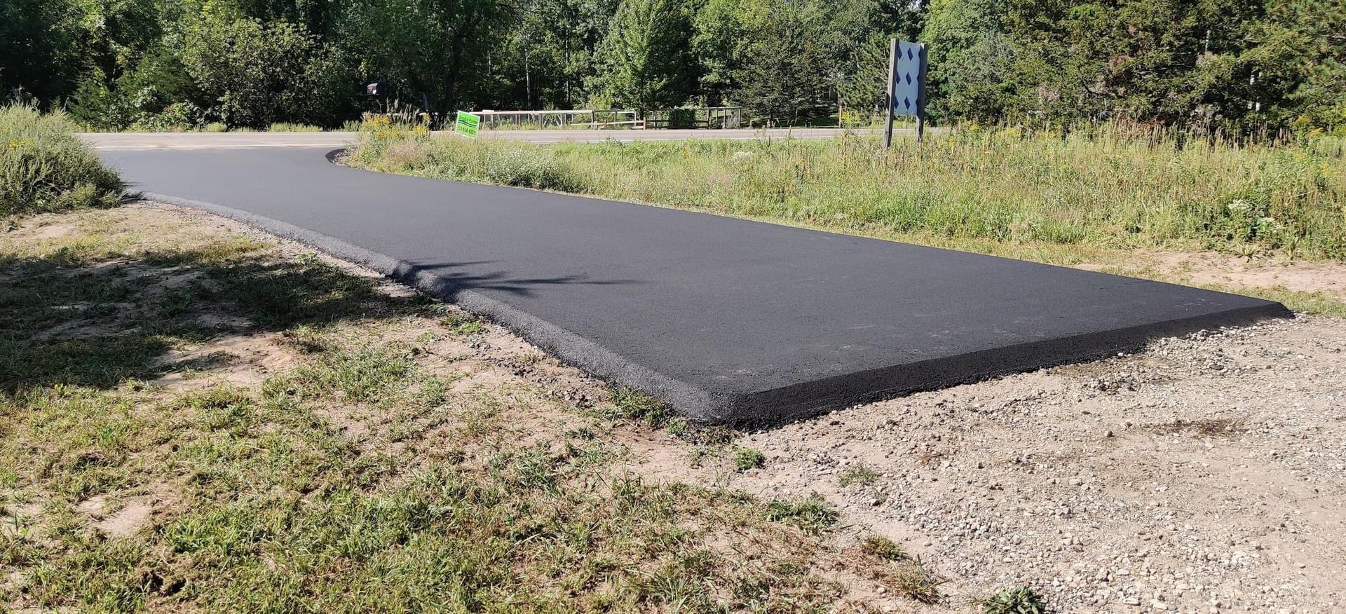 Freshly paved black asphalt path through a grassy, wooded area with a small signpost.