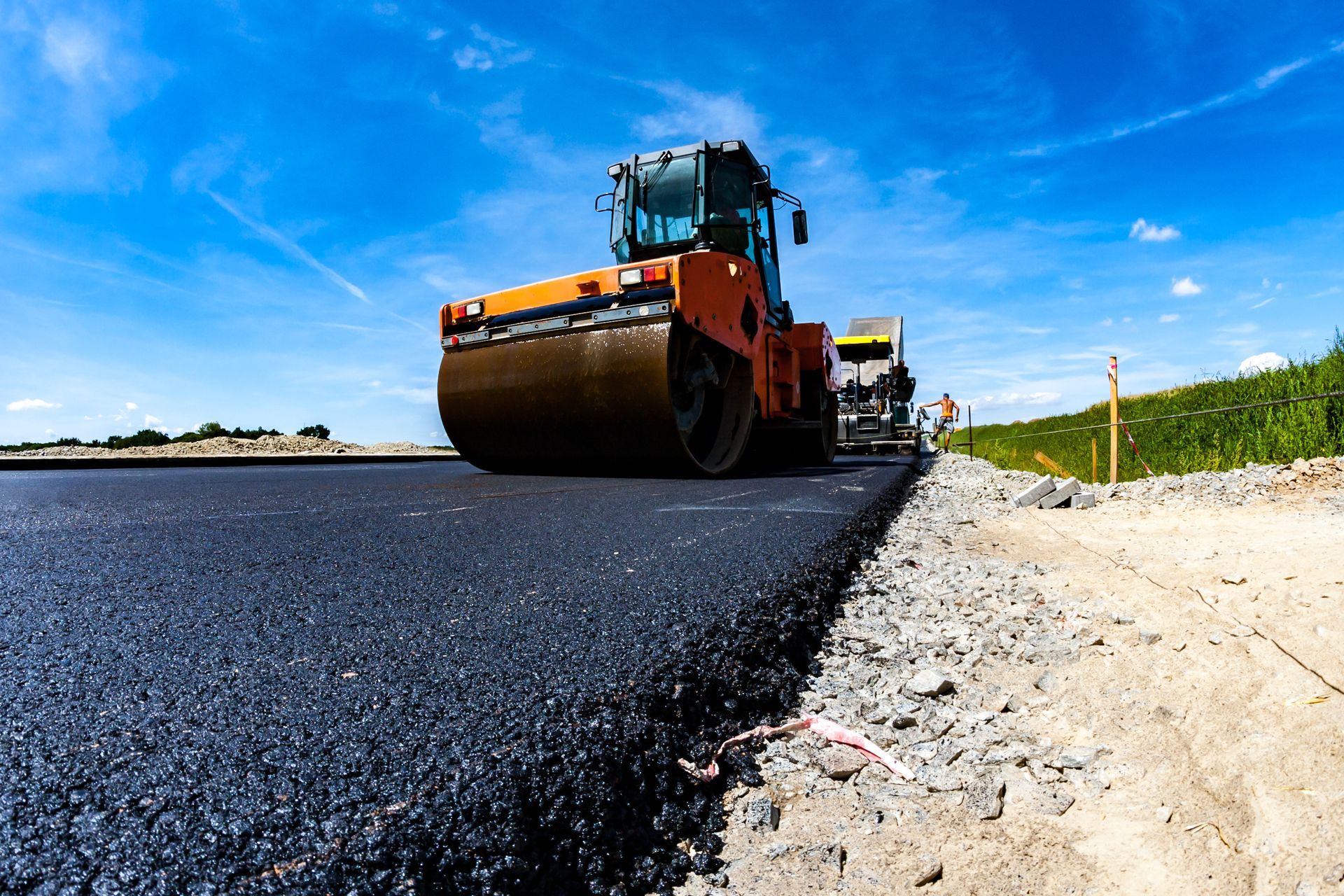 Road roller paving fresh black asphalt beside a sandy shoulder under a bright blue sky