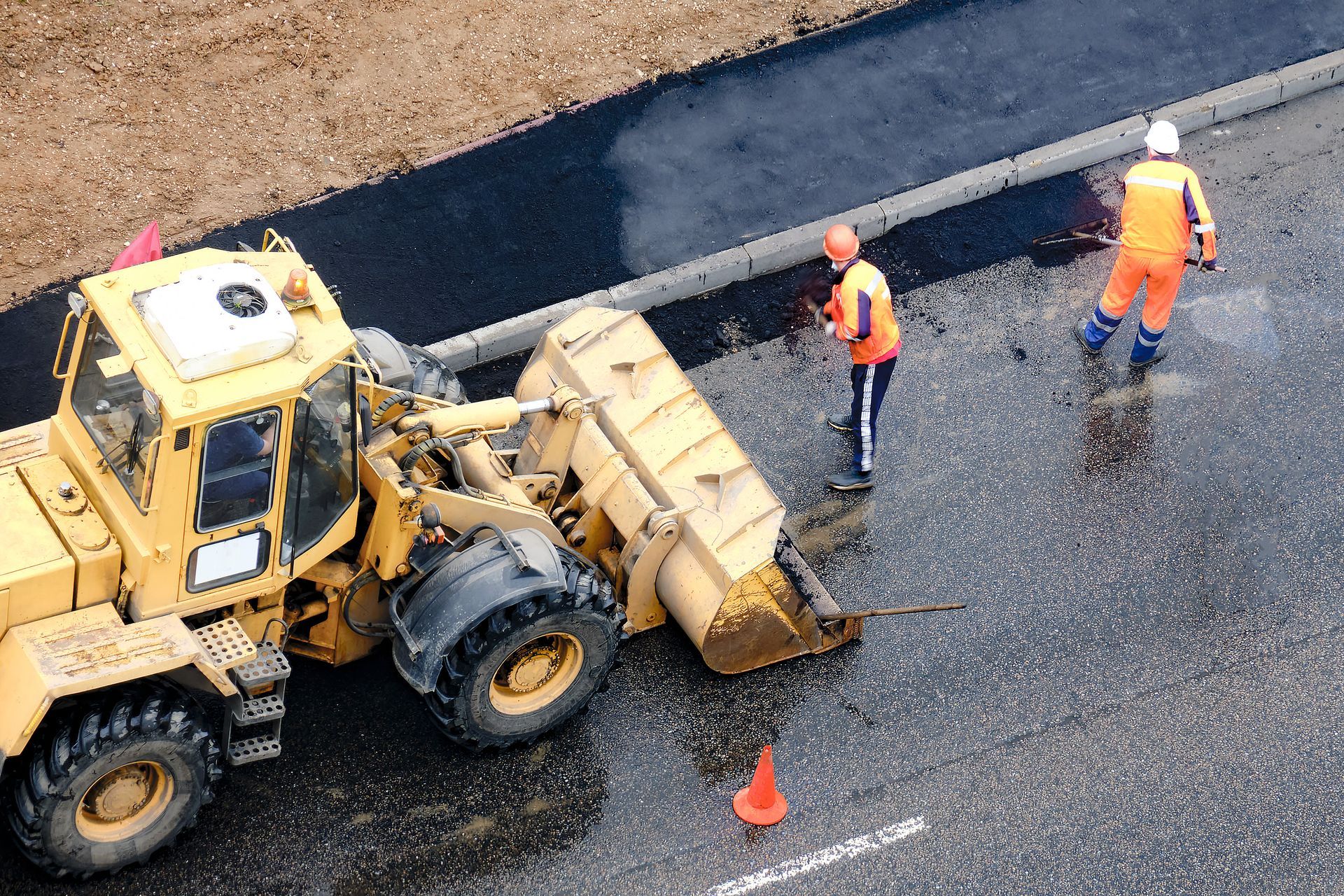 Roadwork crew beside a yellow paving machine on a wet road, with two workers in orange safety gear.