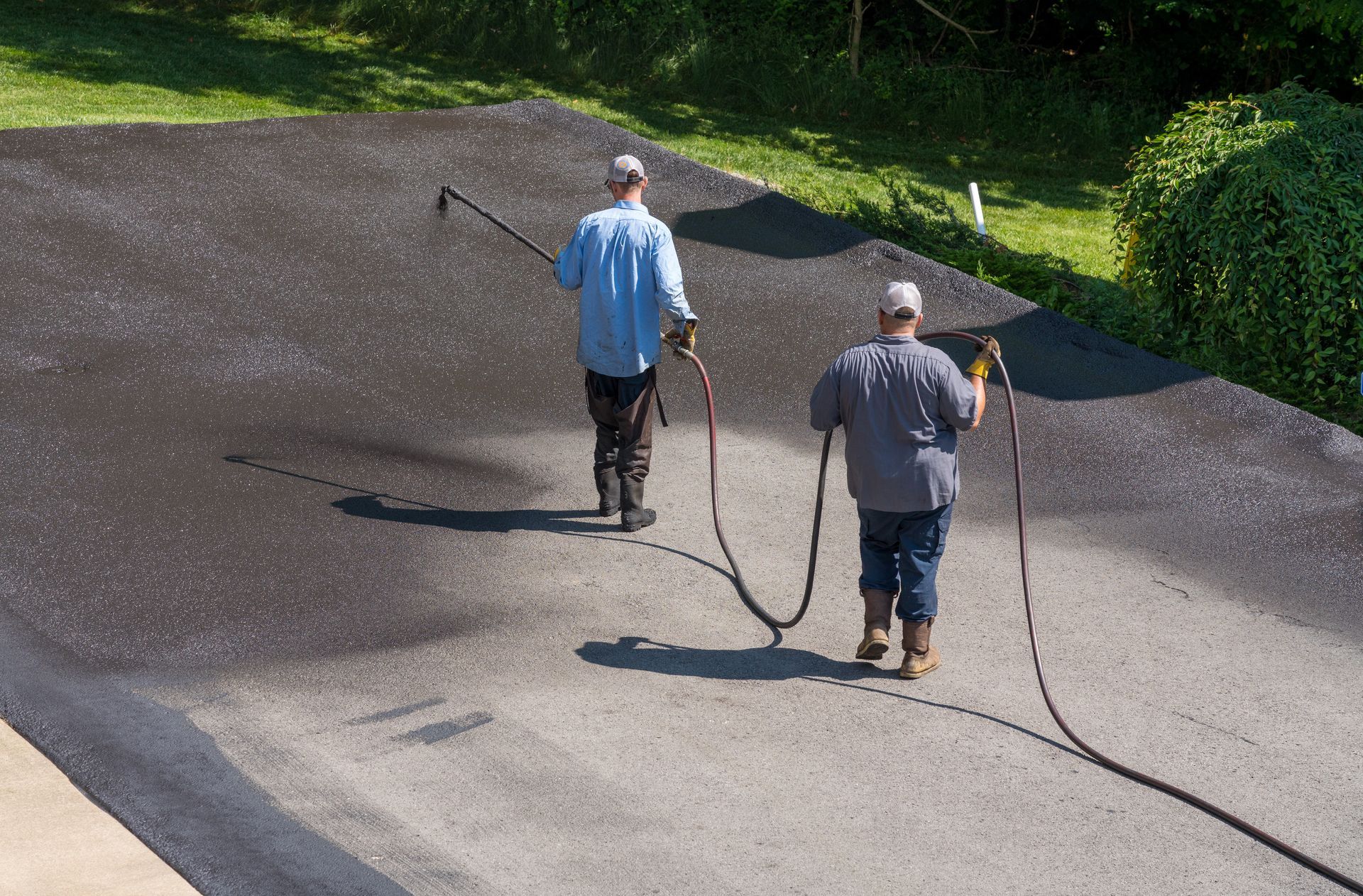 Two workers spraying a dark surface outdoors with hoses, near grass and trees.
