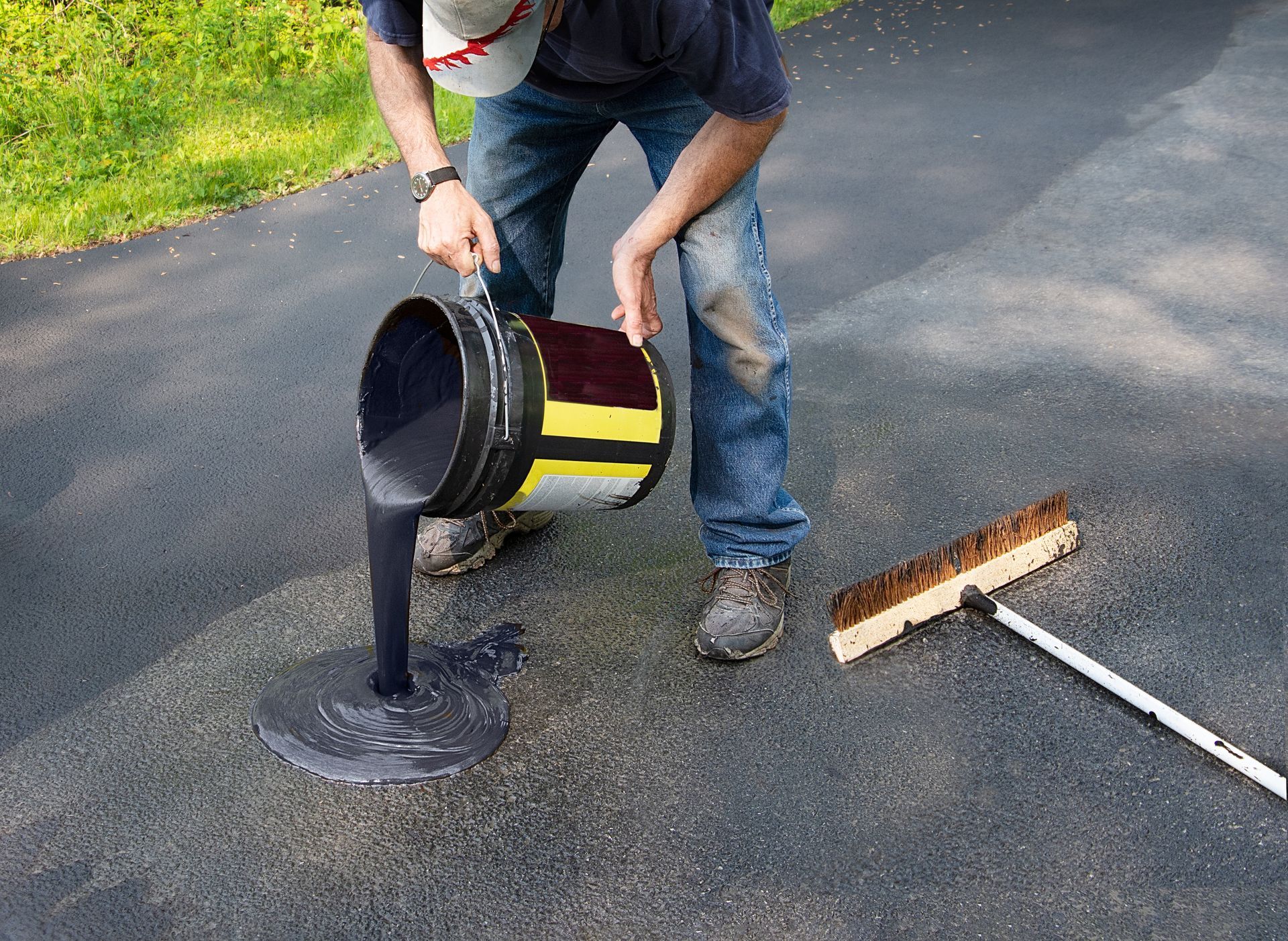 Person pouring black liquid from a bucket onto a driveway beside a broom and squeegee
