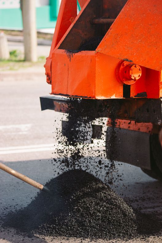 Orange construction vehicle unloading black gravel onto a street.