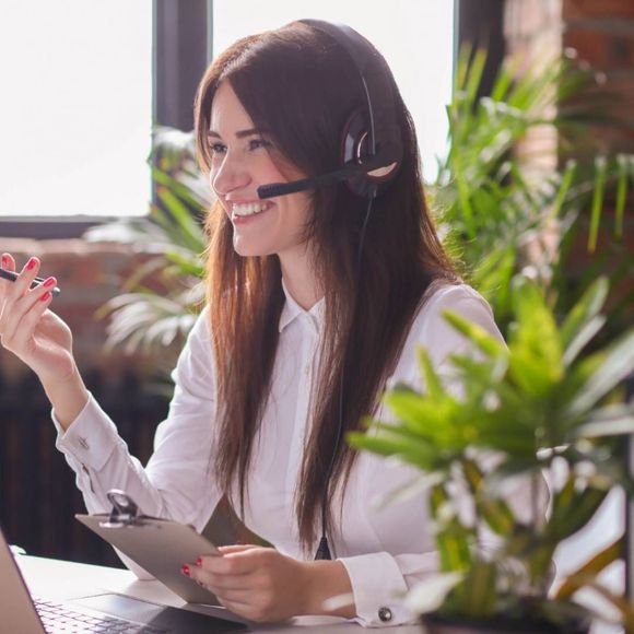 Mujer con auriculares, sonriendo, sosteniendo portapapeles y bolígrafo, en la oficina con plantas.