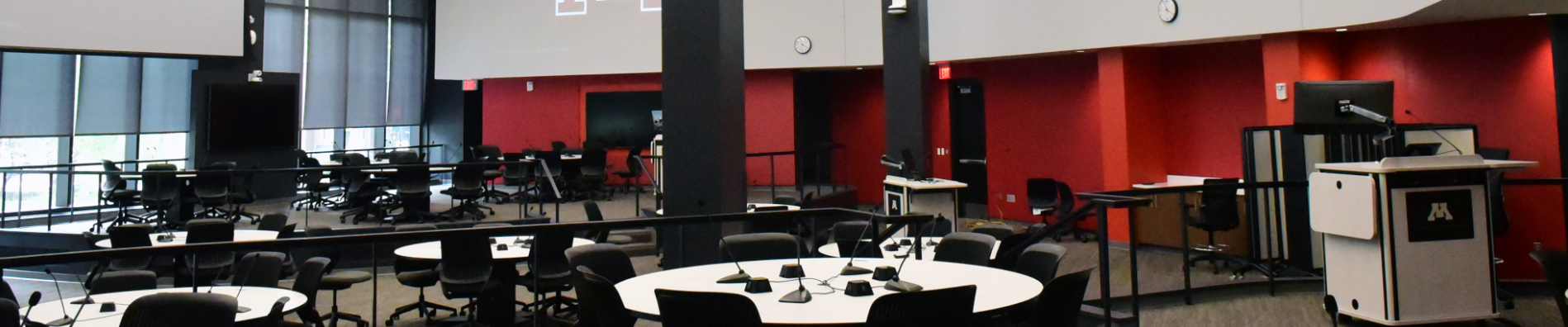 Photograph of the health science classroom at the University of Minnesota, with multiple white round tables, black chairs, and integrated screens, microphones and technology
