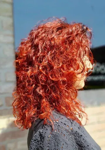 Il parrucchiere spruzza lacca sui capelli di una persona seduta, con capelli lunghi e chiari; l'ambiente è luminoso e circondato da finestre.