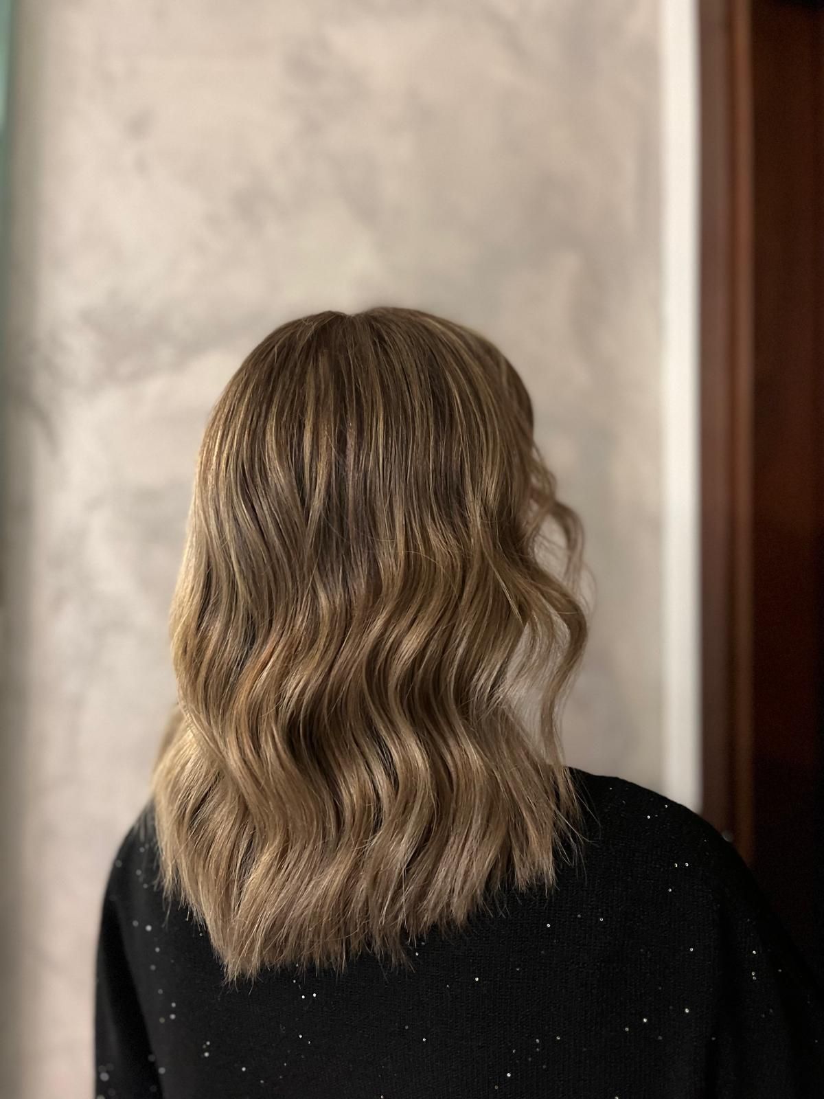 Donna con capelli castani e biondi mossi. Indossa un maglione nero maculato, contro una parete grigia.