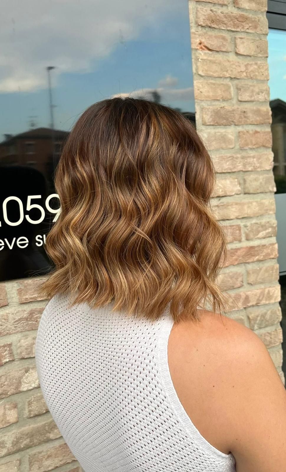 Donna con capelli castani ondulati. I capelli presentano riflessi più chiari sulle punte. In piedi fuori da un edificio in mattoni.