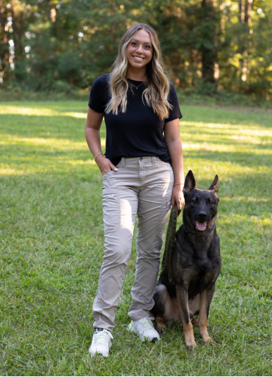 Woman with long blonde hair, black shirt, tan pants, white shoes, and a dog in a grassy setting.