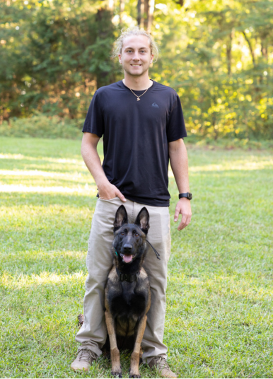 Man stands with a dog, both looking forward, on a grassy lawn with trees in the background.