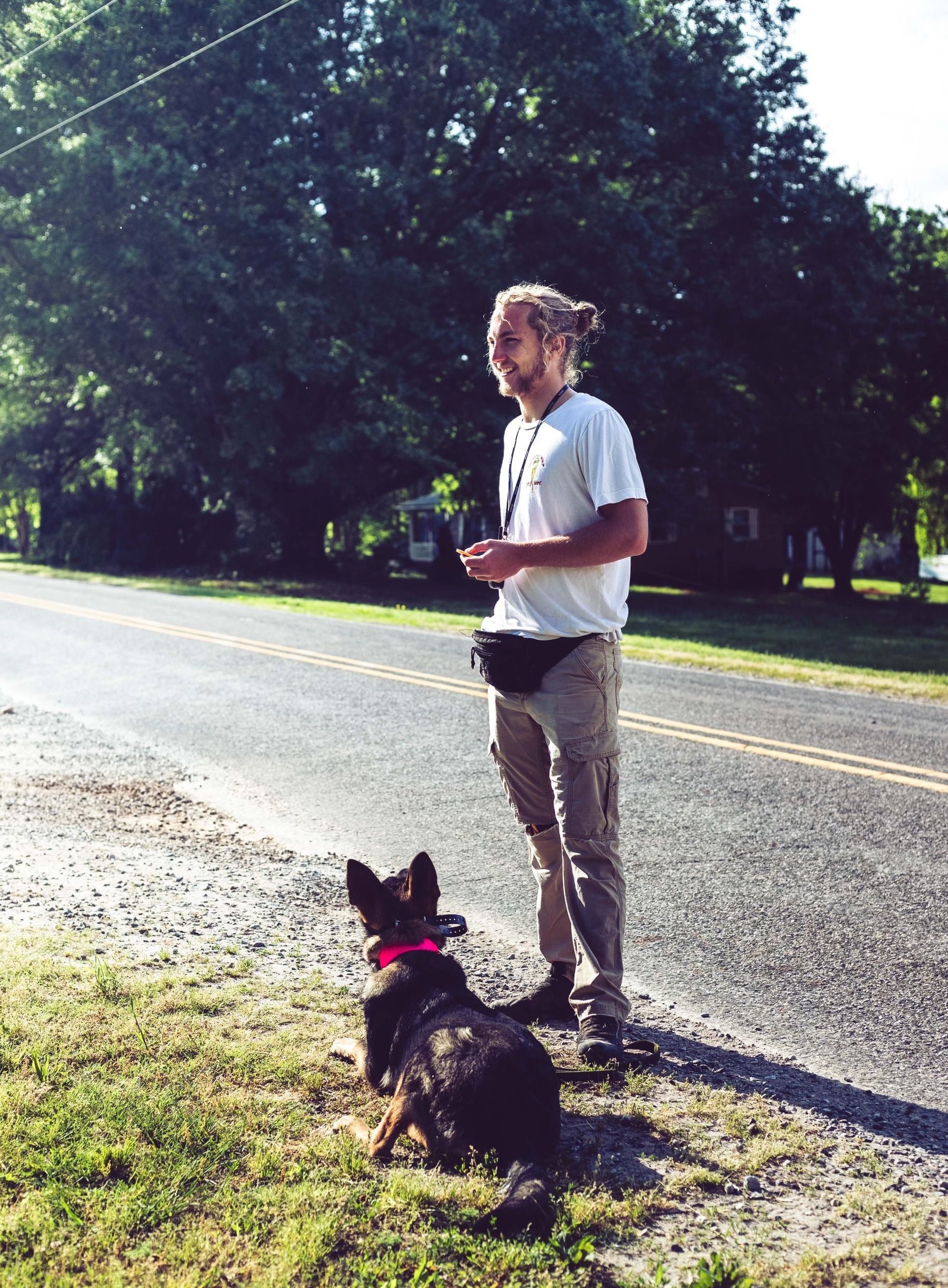 Man in white shirt and tan pants standing on roadside with dog.