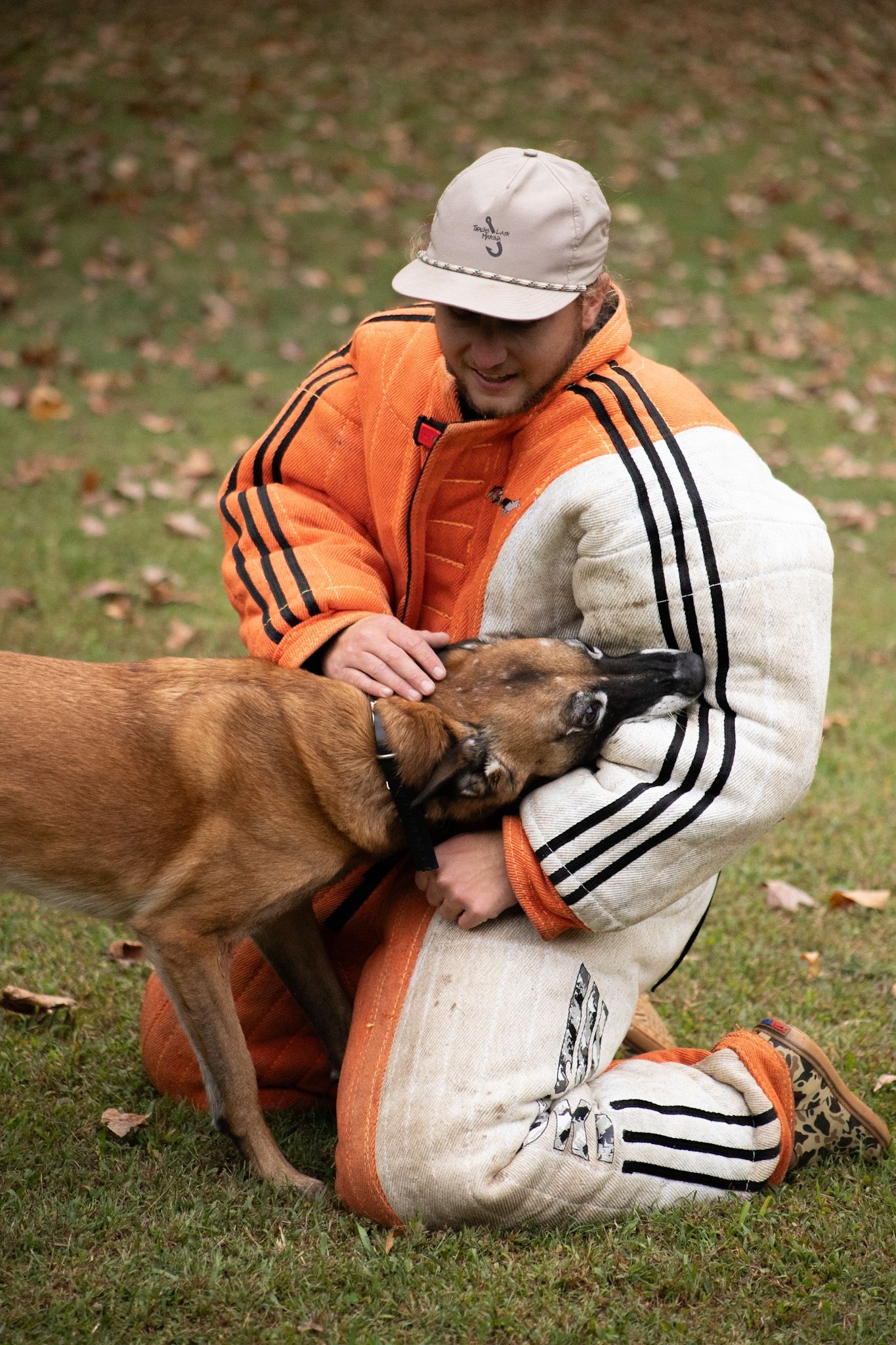 Man in protective gear kneels, petting a working dog on a grassy field.