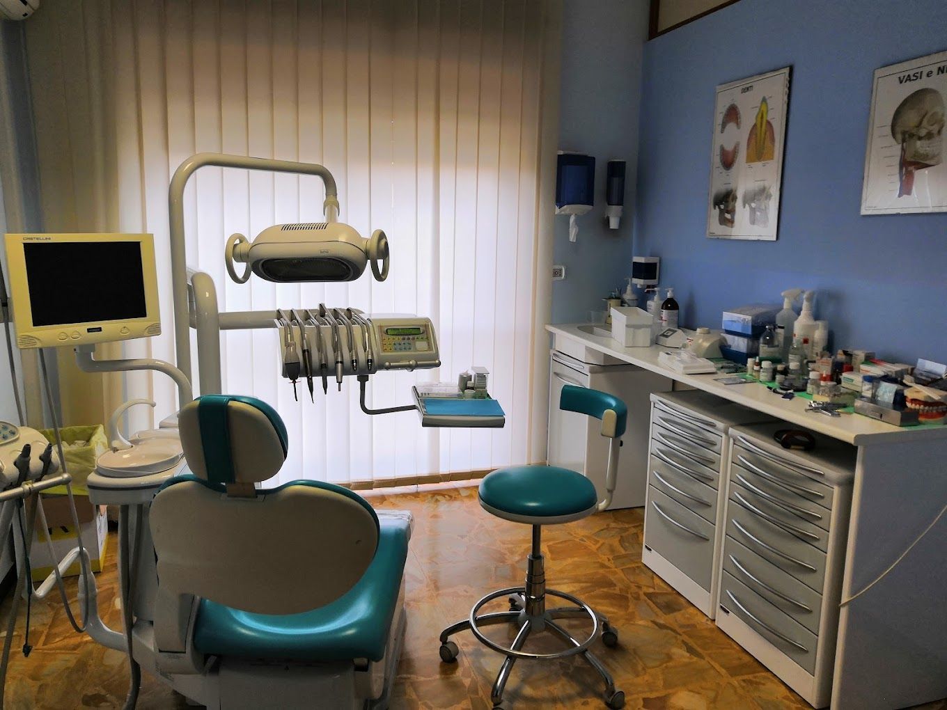 Sala visite odontoiatriche vuota con poltrona, lampada a soffitto, monitor e armadietti su pareti blu.