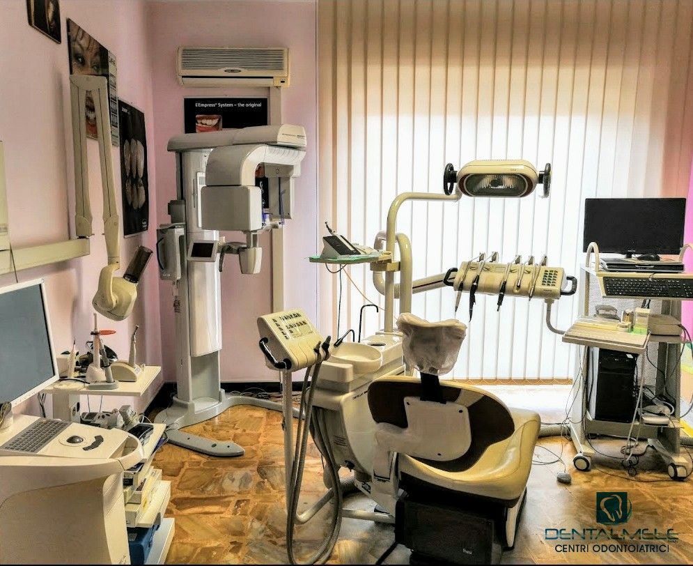 Studio dentistico con poltrona per la visita, apparecchio radiografico, monitor e luci a soffitto vicino a una finestra.