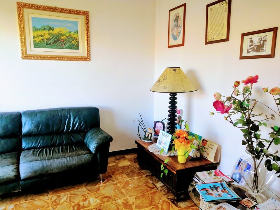 Soggiorno con divano verde scuro, lampada, quadri alle pareti, fiori e foto incorniciate su un tavolino.