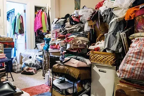 Cluttered room filled with piles of clothes, bags, and items. A closet with hanging clothes is visible.