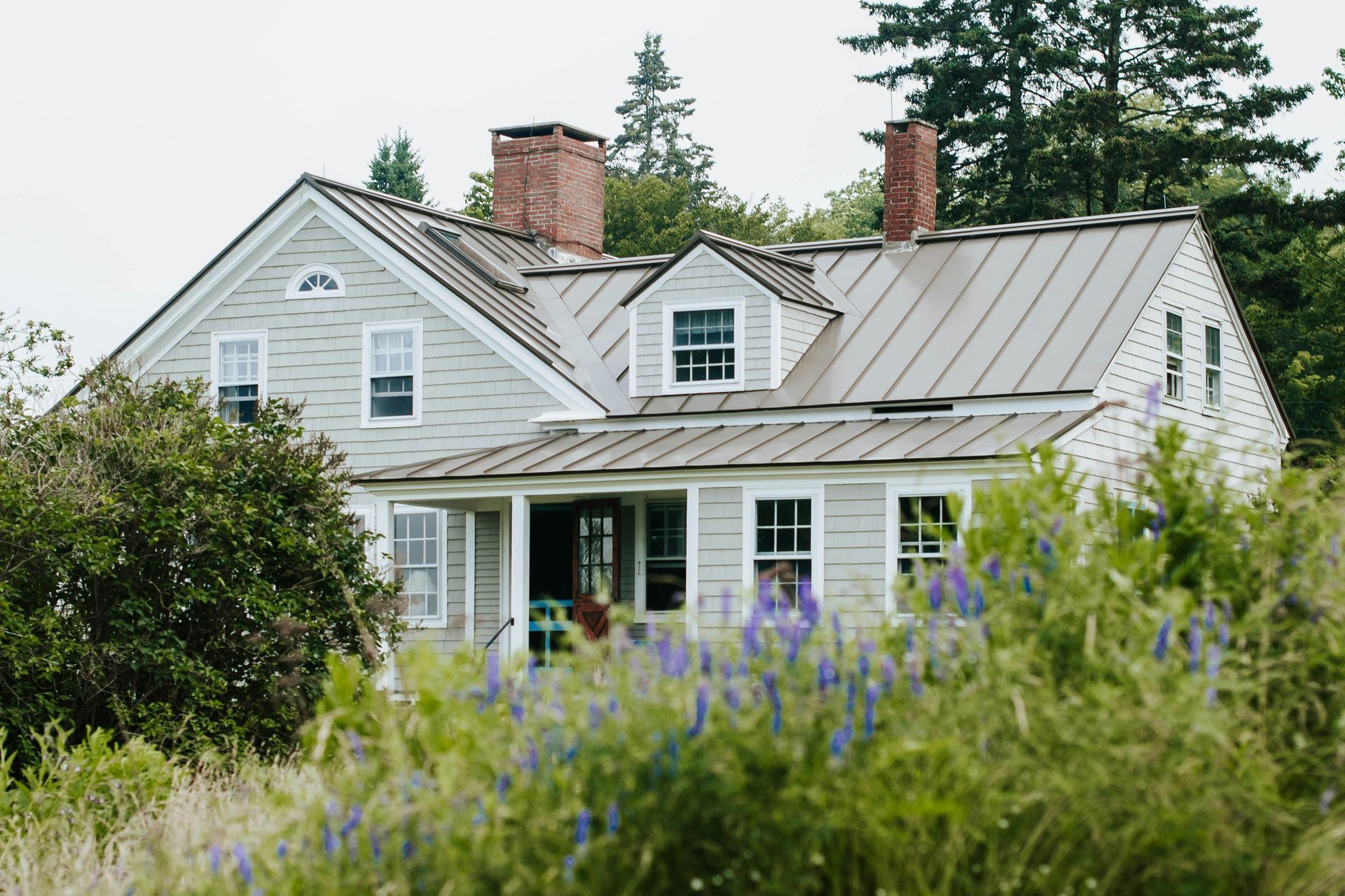 A white house with a metal roof is surrounded by trees and flowers.