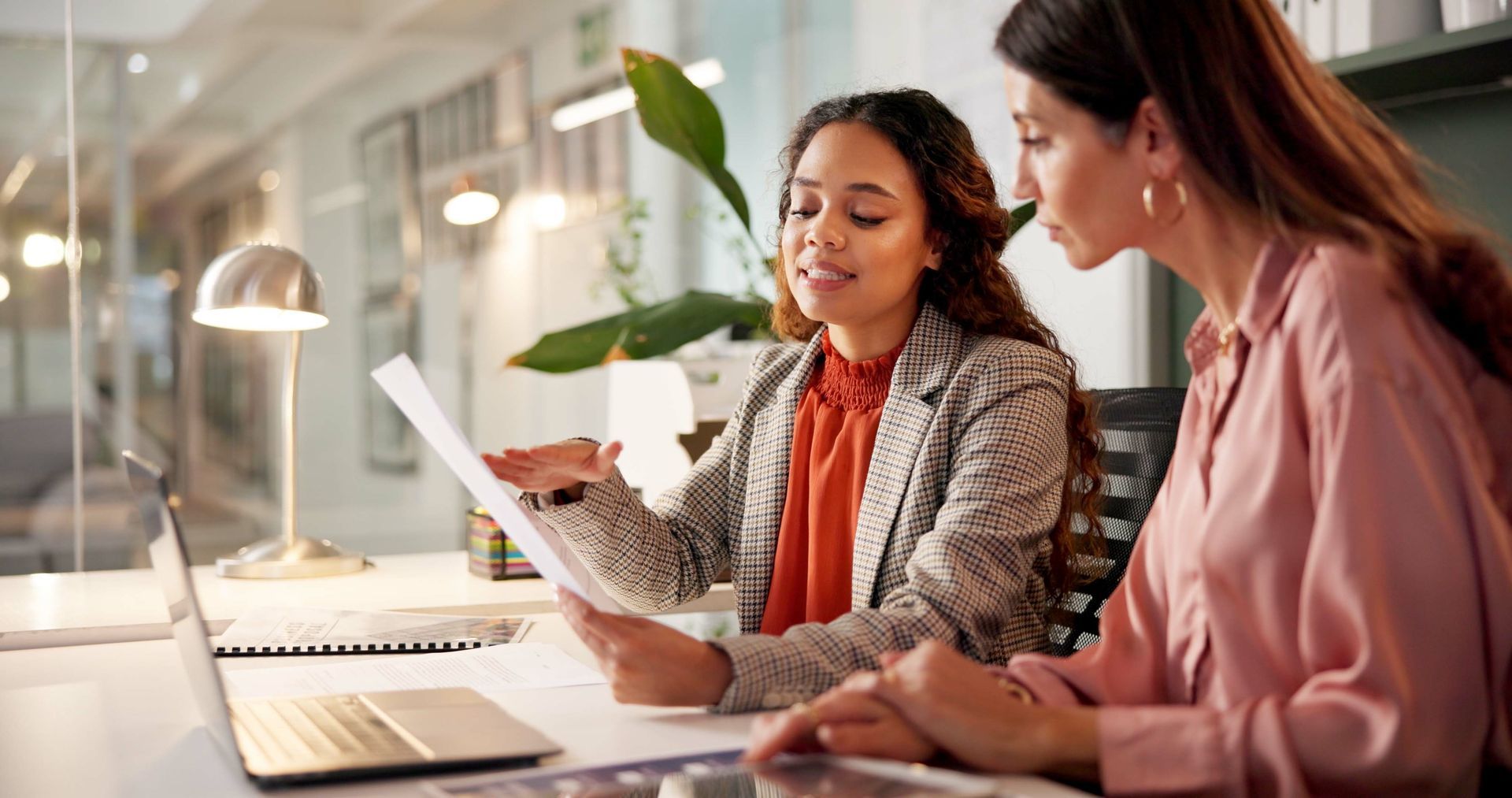 Two women reviewing documents at a desk in an office. One points at the paper, and both look focused. Two women reviewing documents at a desk in an office. One points at the paper, and both look focused.