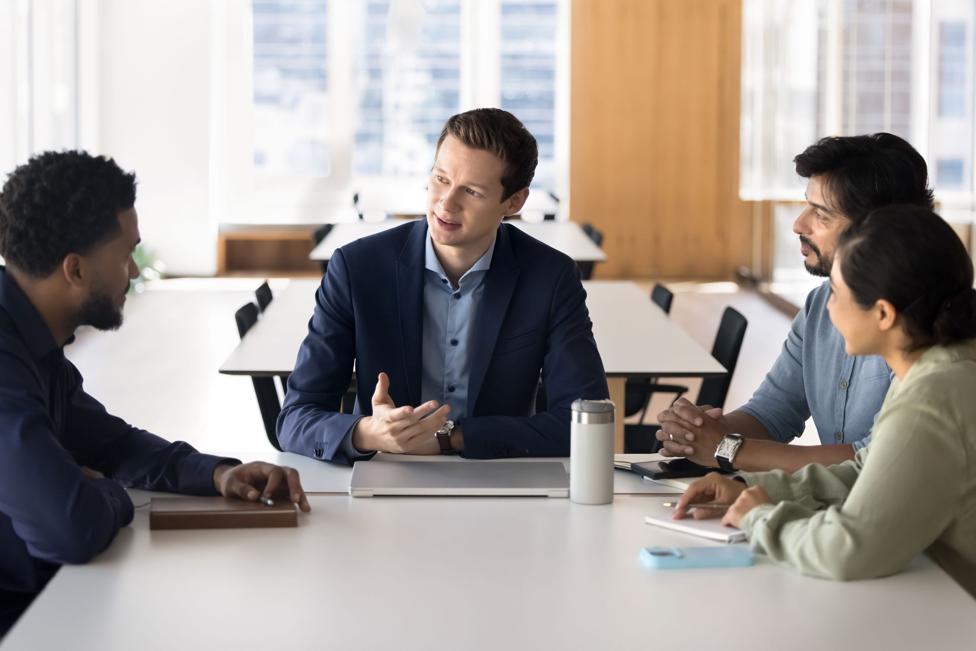 Four people in a meeting, centered around a table, one man gesturing. Bright, modern office setting. Four people in a meeting, centered around a table, one man gesturing. Bright, modern office setting.