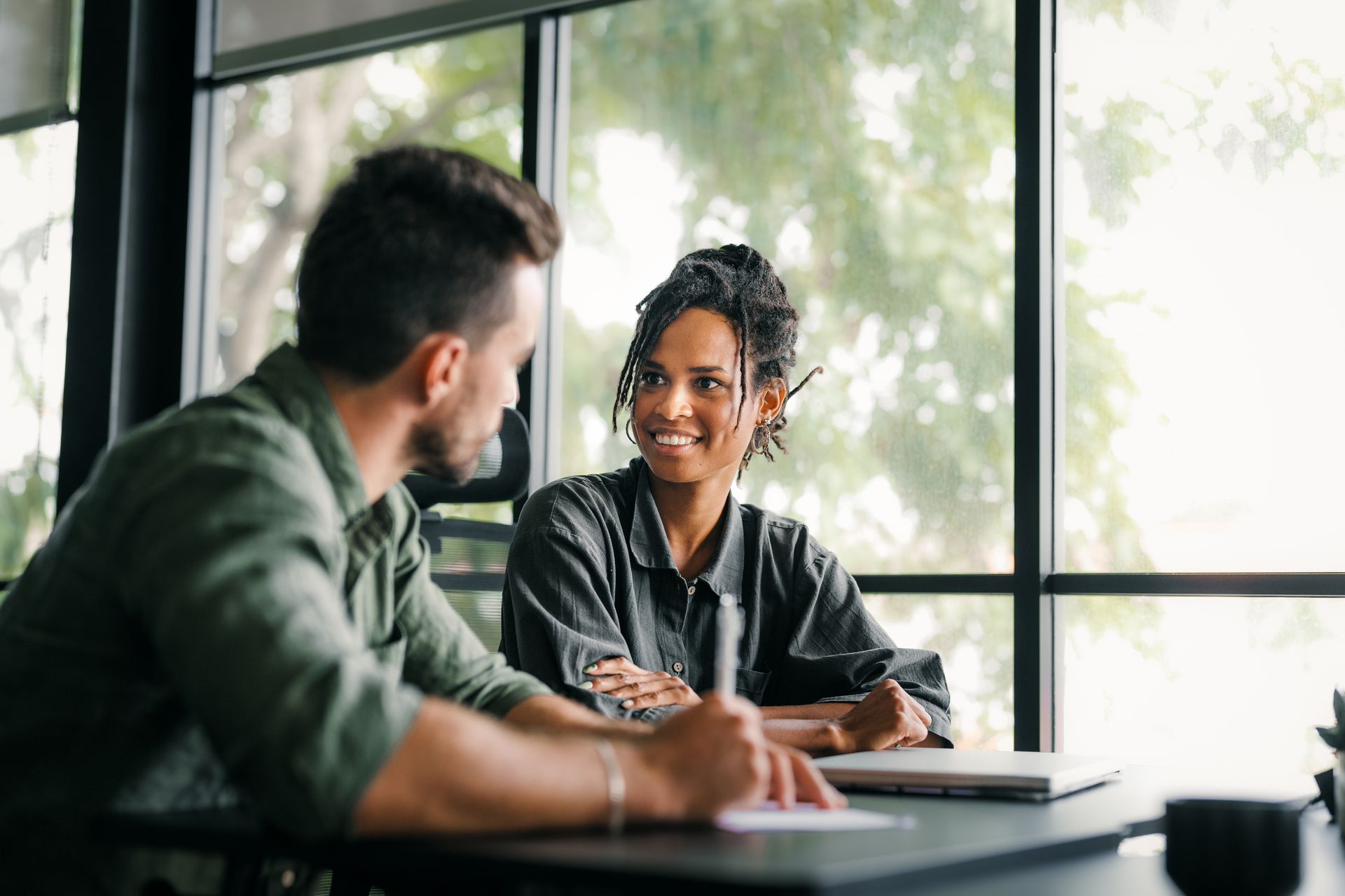 Two people in a discussion at a table near a window. Woman smiles while looking at the man writing on a document. Two people in a discussion at a table near a window. Woman smiles while looking at the man writing on a document.