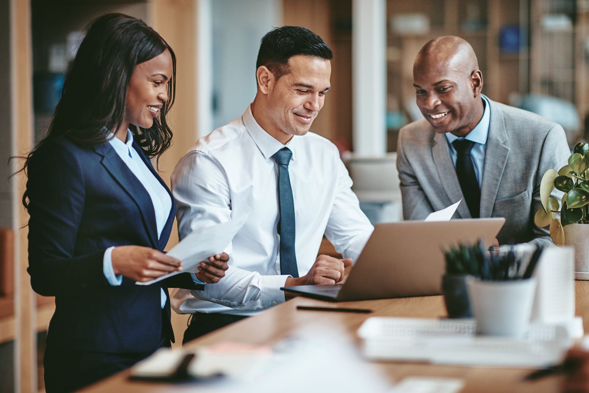 Three people in business attire collaborate around a laptop, smiling in an office setting. Three people in business attire collaborate around a laptop, smiling in an office setting.