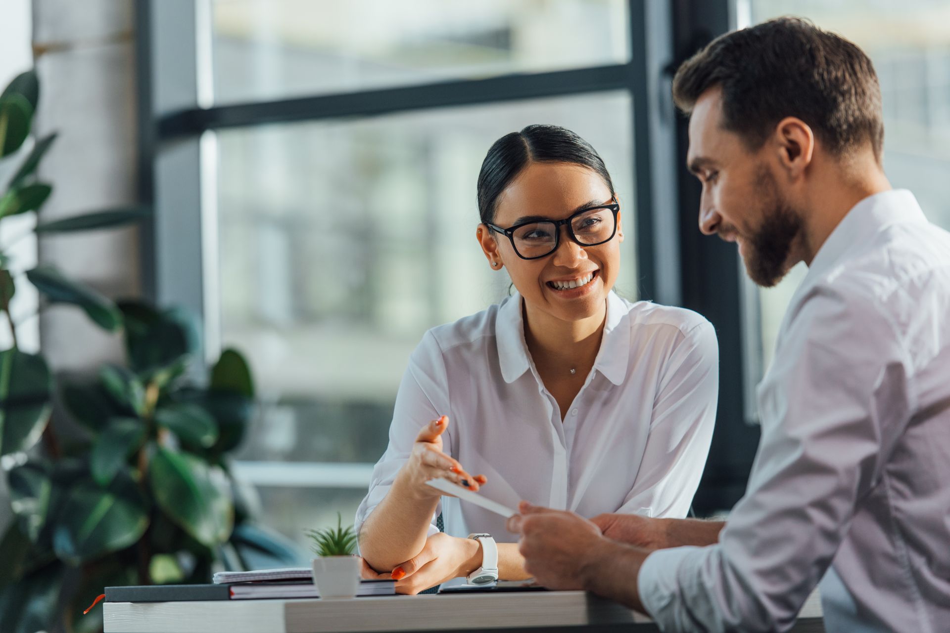Two people in a bright office: woman with glasses smiles, gesturing at a paper; man looks at the paper and smiles. Two people in a bright office: woman with glasses smiles, gesturing at a paper; man looks at the paper and smiles.