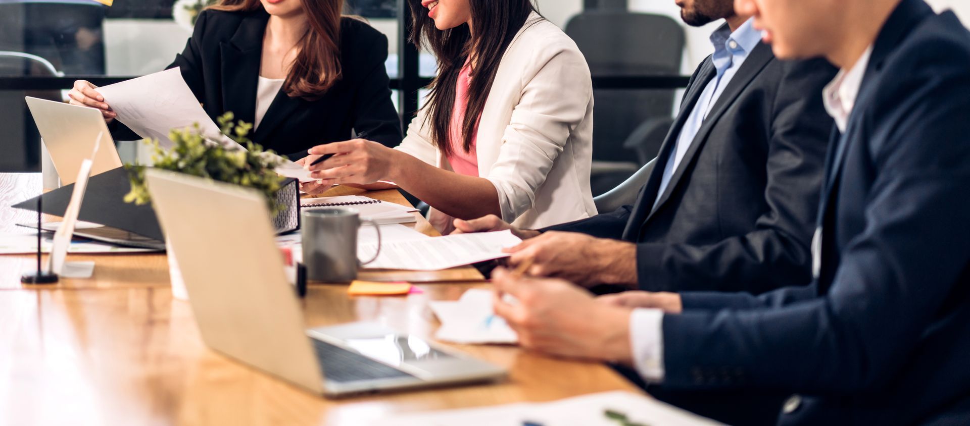 People in business attire at a conference table with laptops and papers, discussing work. People in business attire at a conference table with laptops and papers, discussing work.