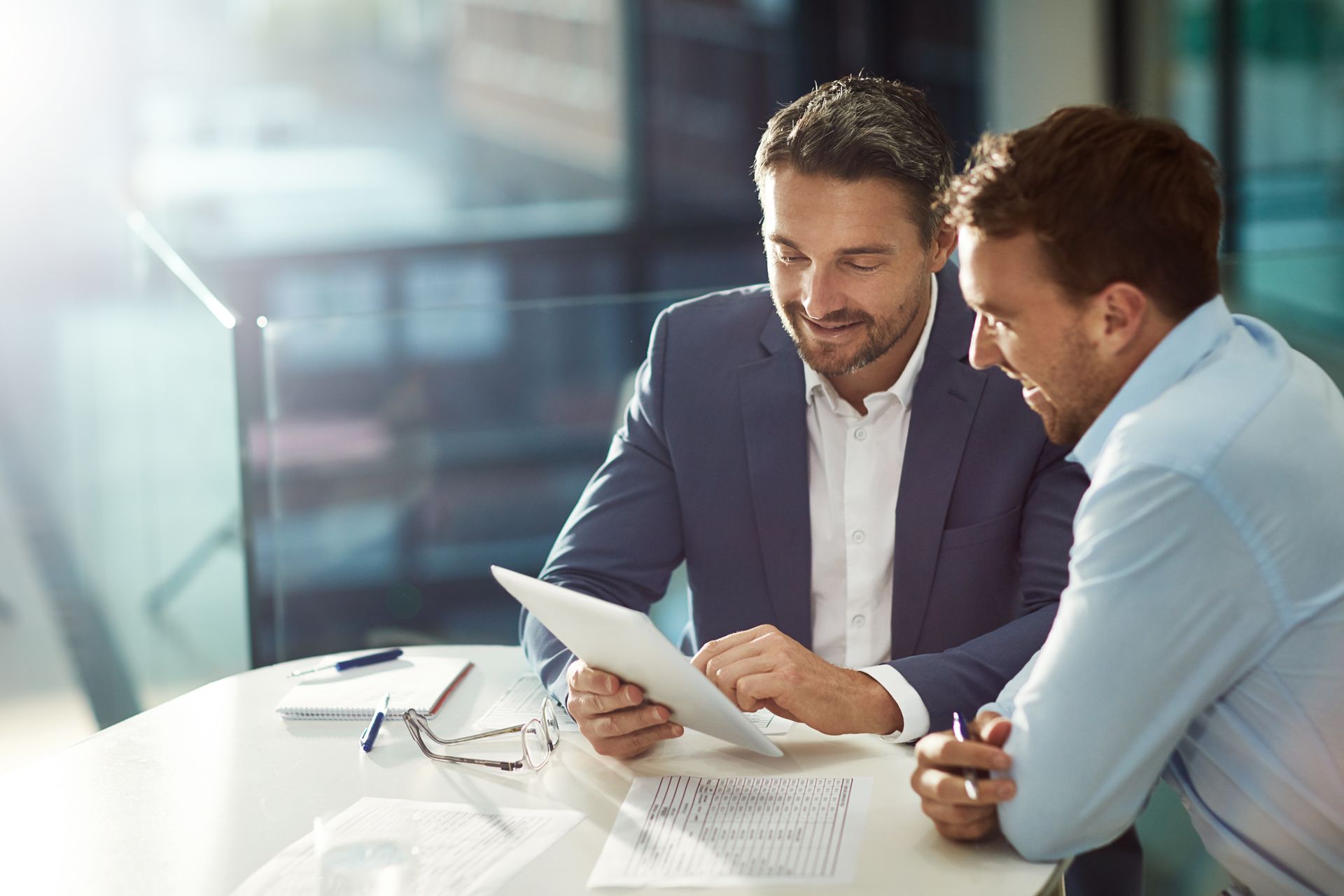 Two men in business attire review a tablet at a table in an office. Two men in business attire review a tablet at a table in an office.