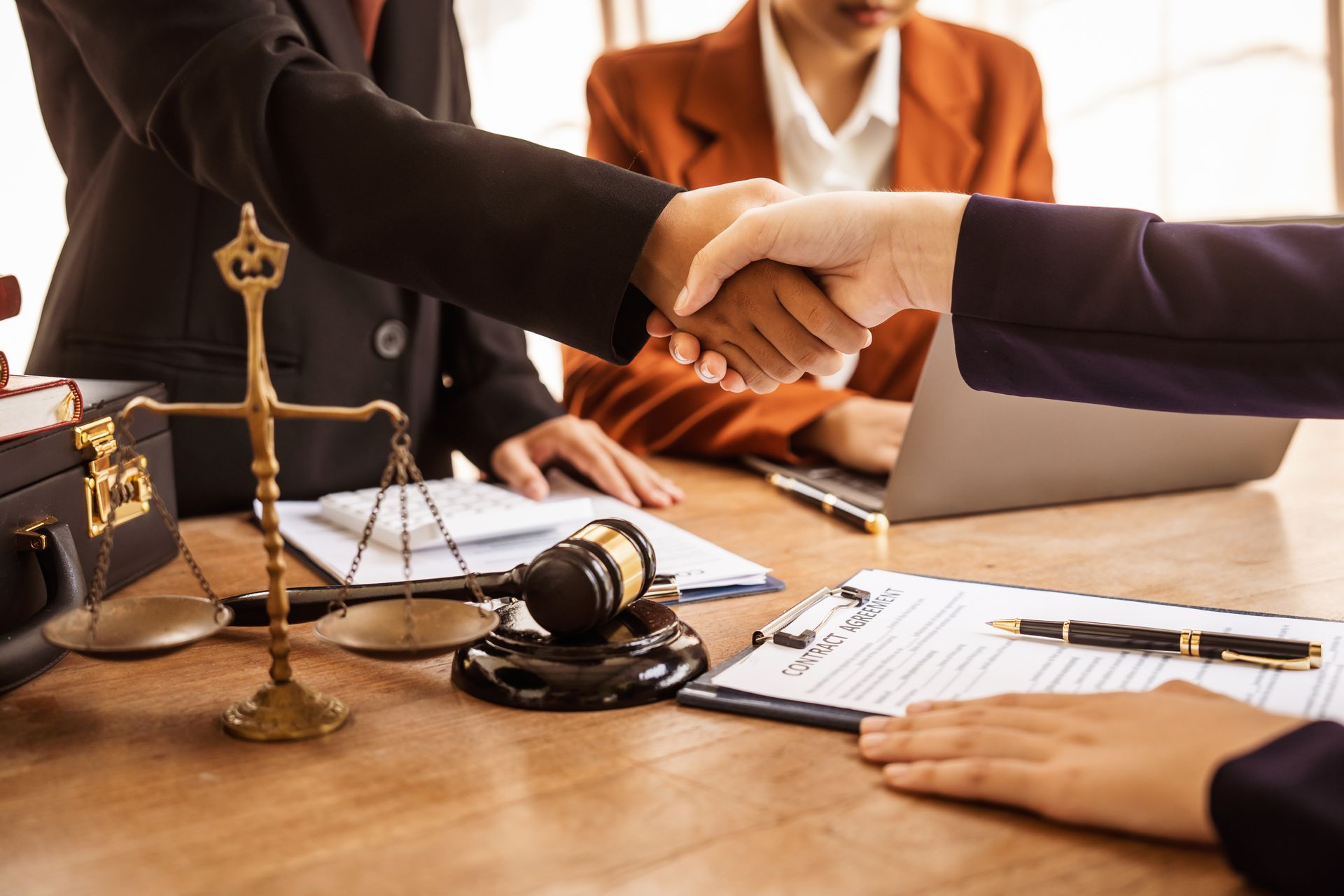 Two people shaking hands over legal documents with a gavel and scales on desk.