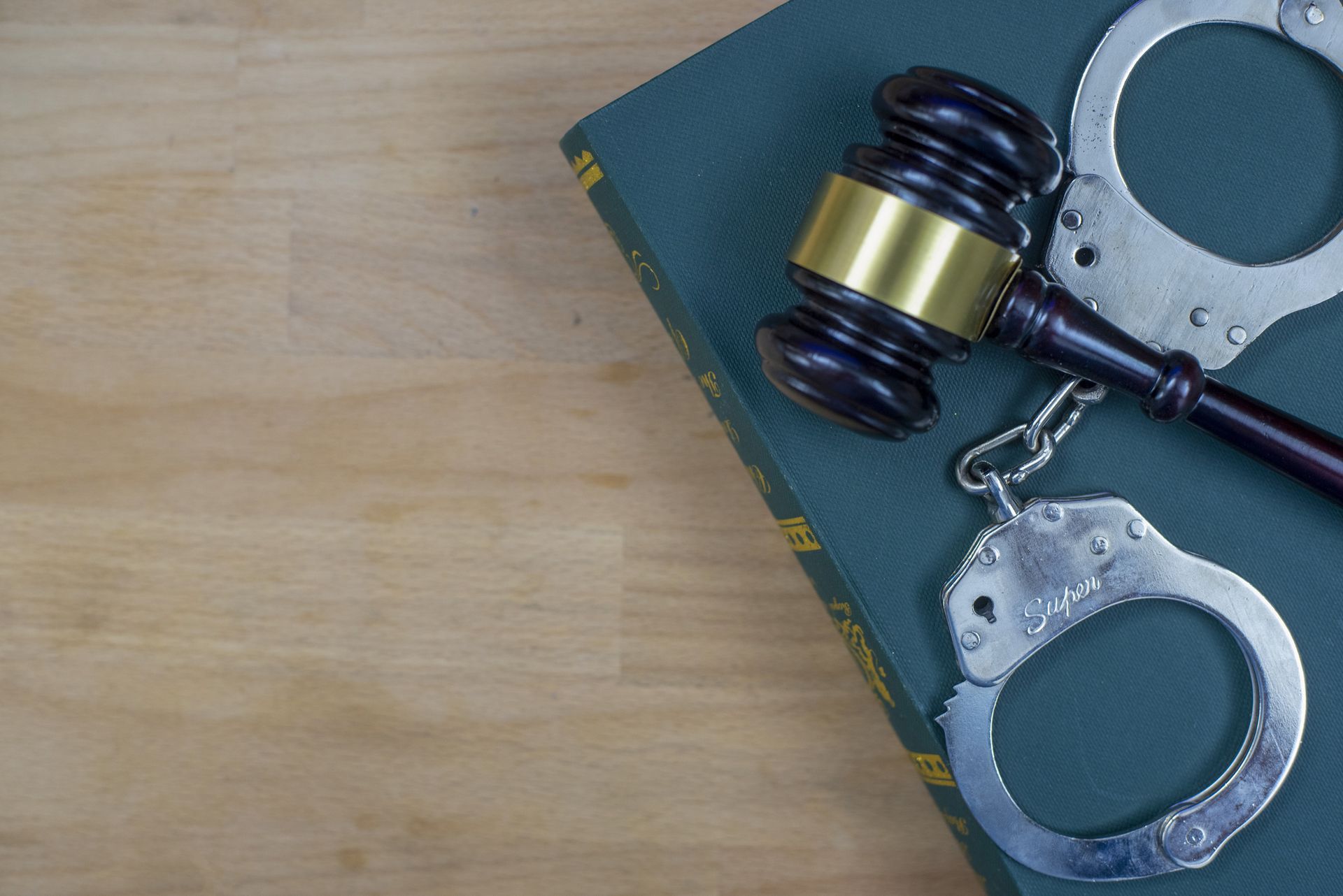 Judge’s gavel, open handcuffs, and green law book placed on wooden surface in legal setting.