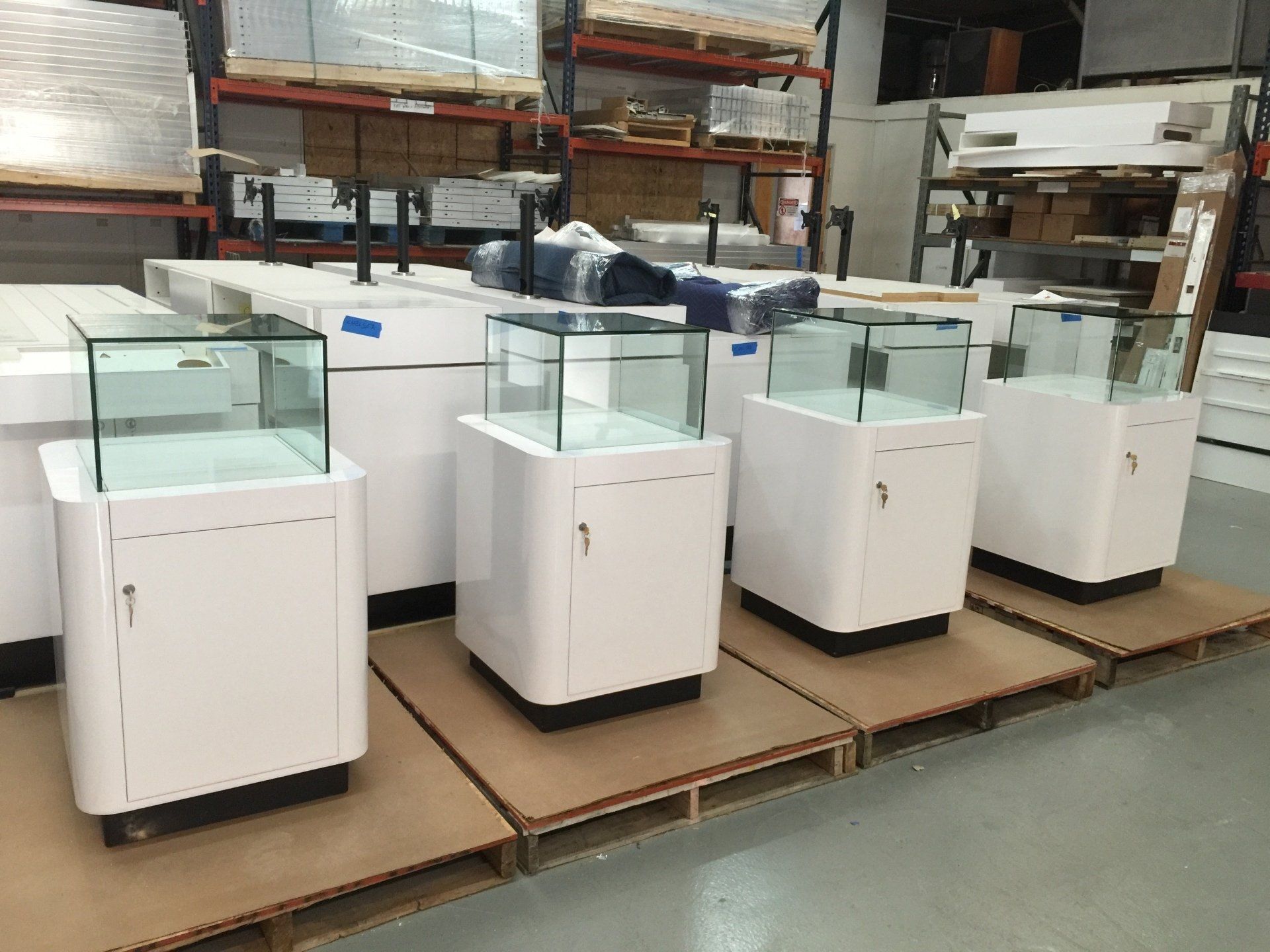 A row of white cabinets with glass doors are sitting on cardboard pallets in a warehouse.