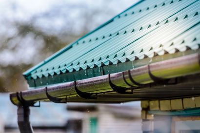 Green corrugated metal roof with rain pouring into a black gutter.