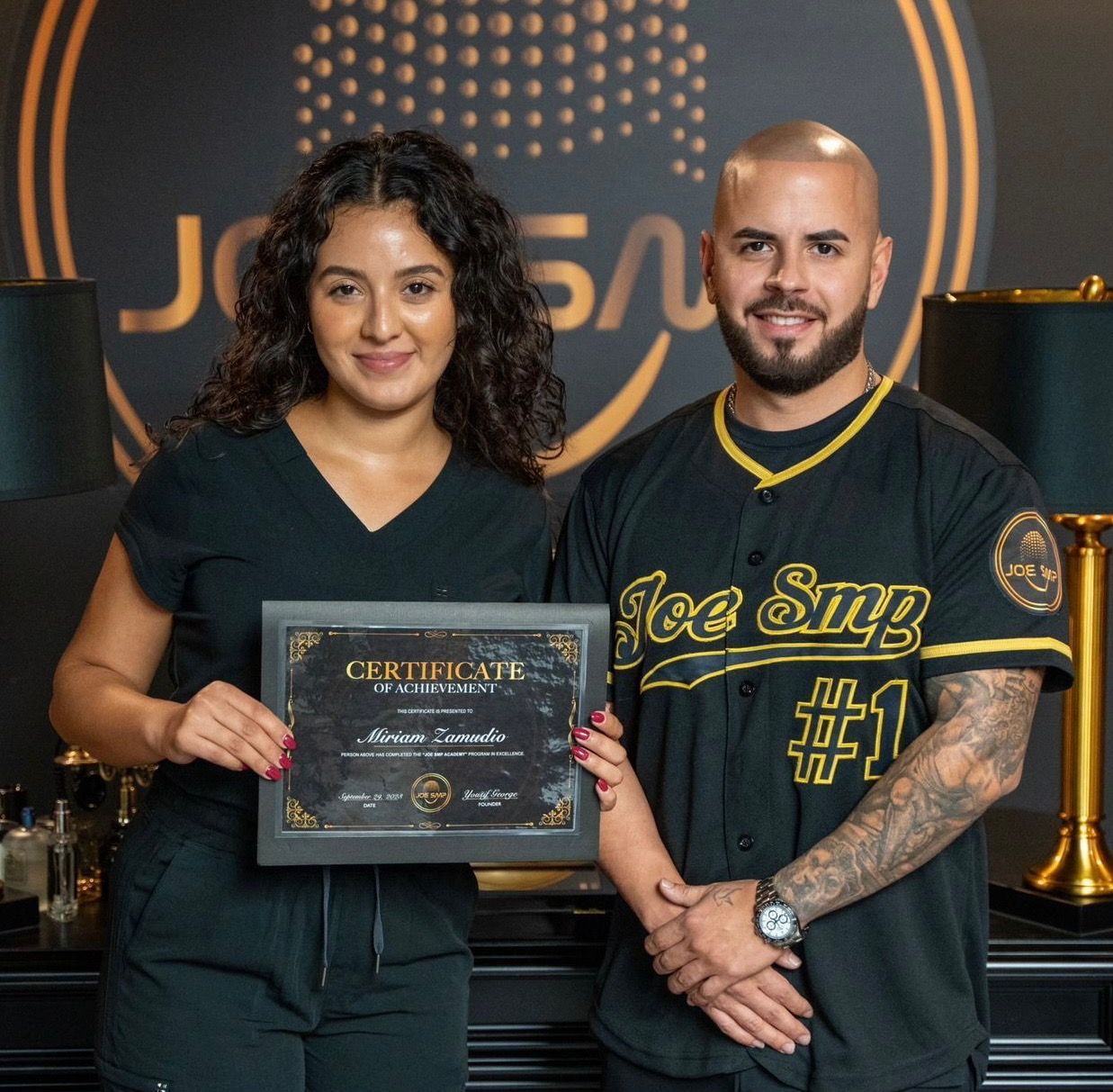 Woman and man holding a certificate in front of a salon logo, both smiling. The woman holds it; the man wears a baseball jersey.