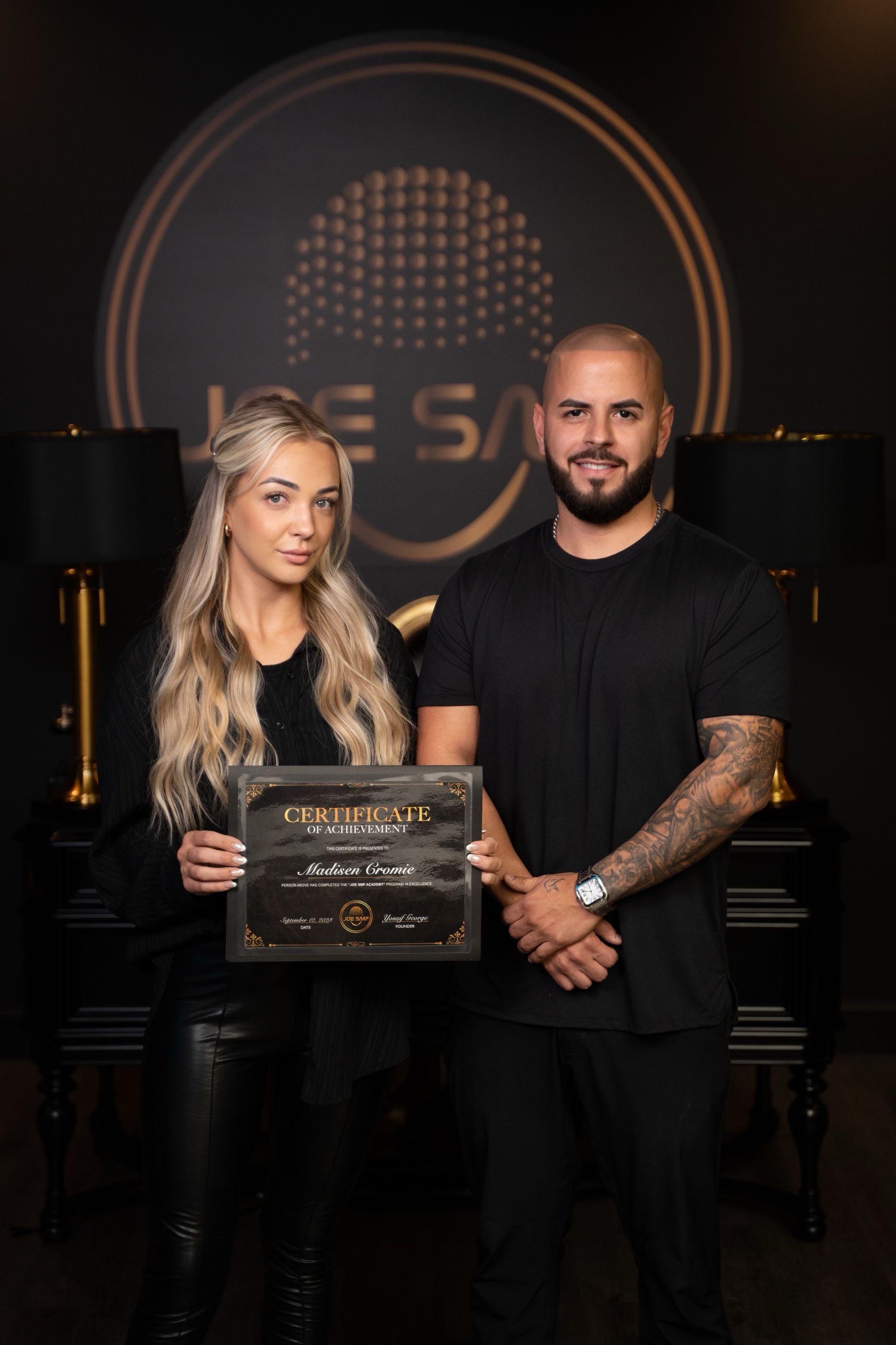 Two people posing, holding a certificate. Man has tattoos, black outfit; woman has blonde hair, leather pants, black top. Dark room, logo in background.