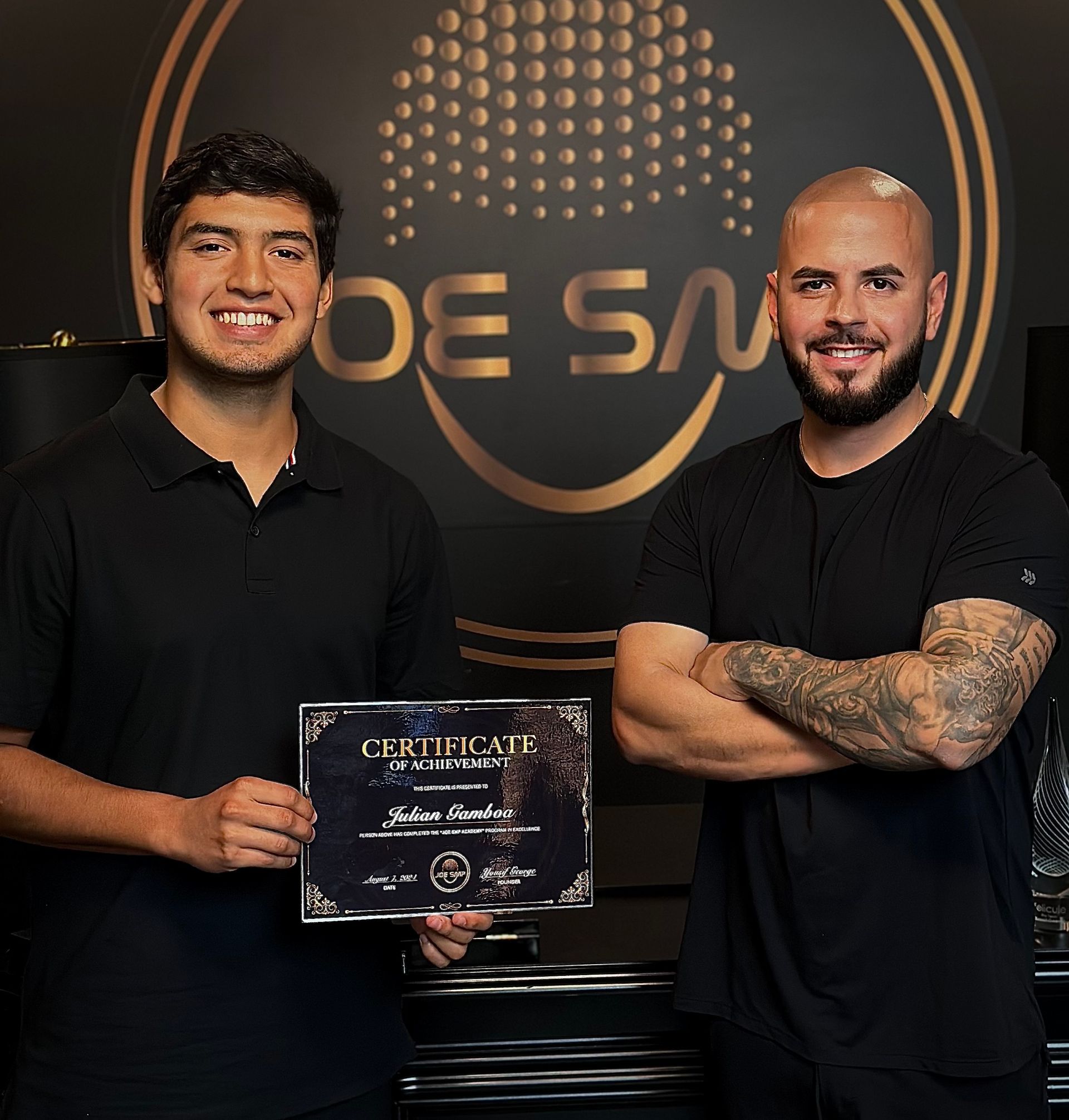 Two men in black, one holding a certificate, smiling.  Behind them is a gold logo: 