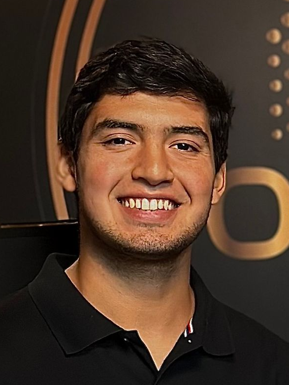 Smiling man in a black polo shirt, with dark hair, indoors in front of a blurred gold background.