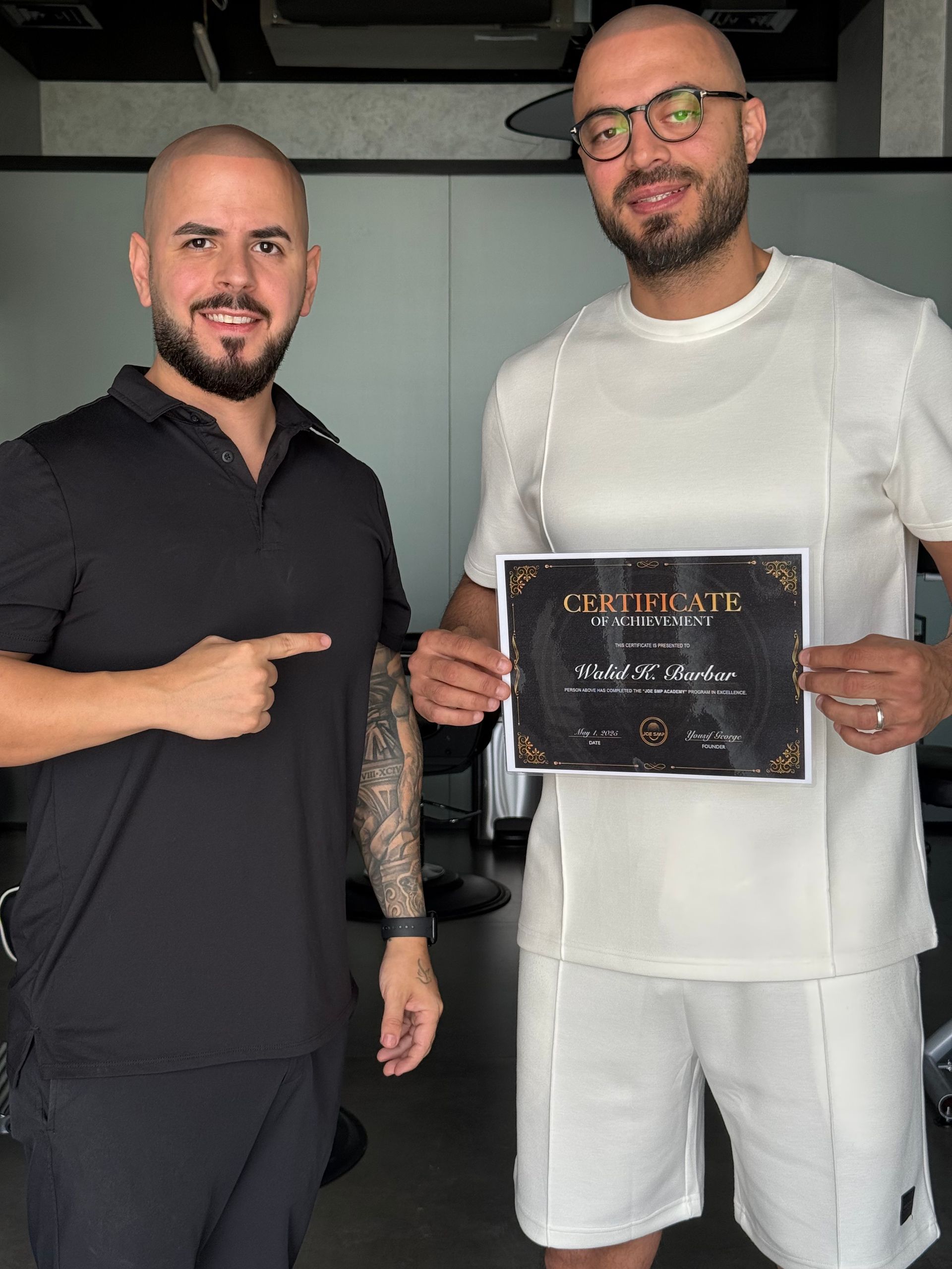 Two men hold a certificate. One points at it, both smiling. They are in a well-lit, professional setting.