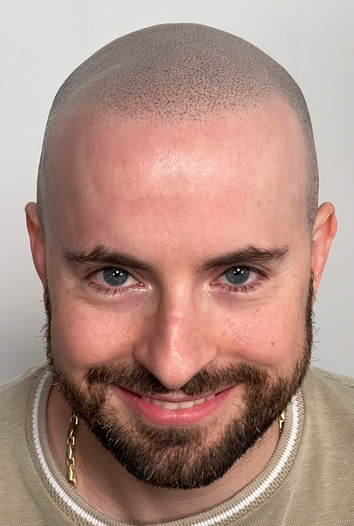 Man with a shaved head and beard, smiling. He's wearing a gold chain and beige shirt.