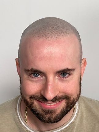 Man with buzzed hair and beard smiling, wearing a beige sweater, against a white backdrop.
