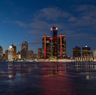 Detroit skyline at dusk with reflections in the water; orange and red lights illuminate buildings.