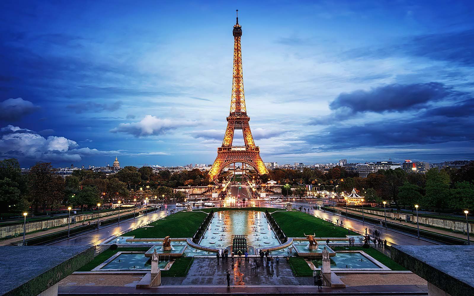 Eiffel Tower at dusk, lit up against a blue sky, reflecting in a water feature. Paris, France.