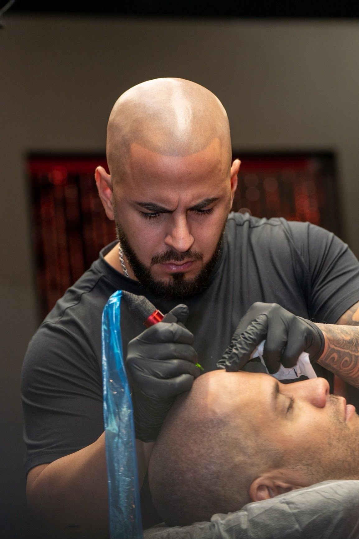 Tattoo artist in black gloves, focused, using a machine on a client's head. Studio setting with red in background.