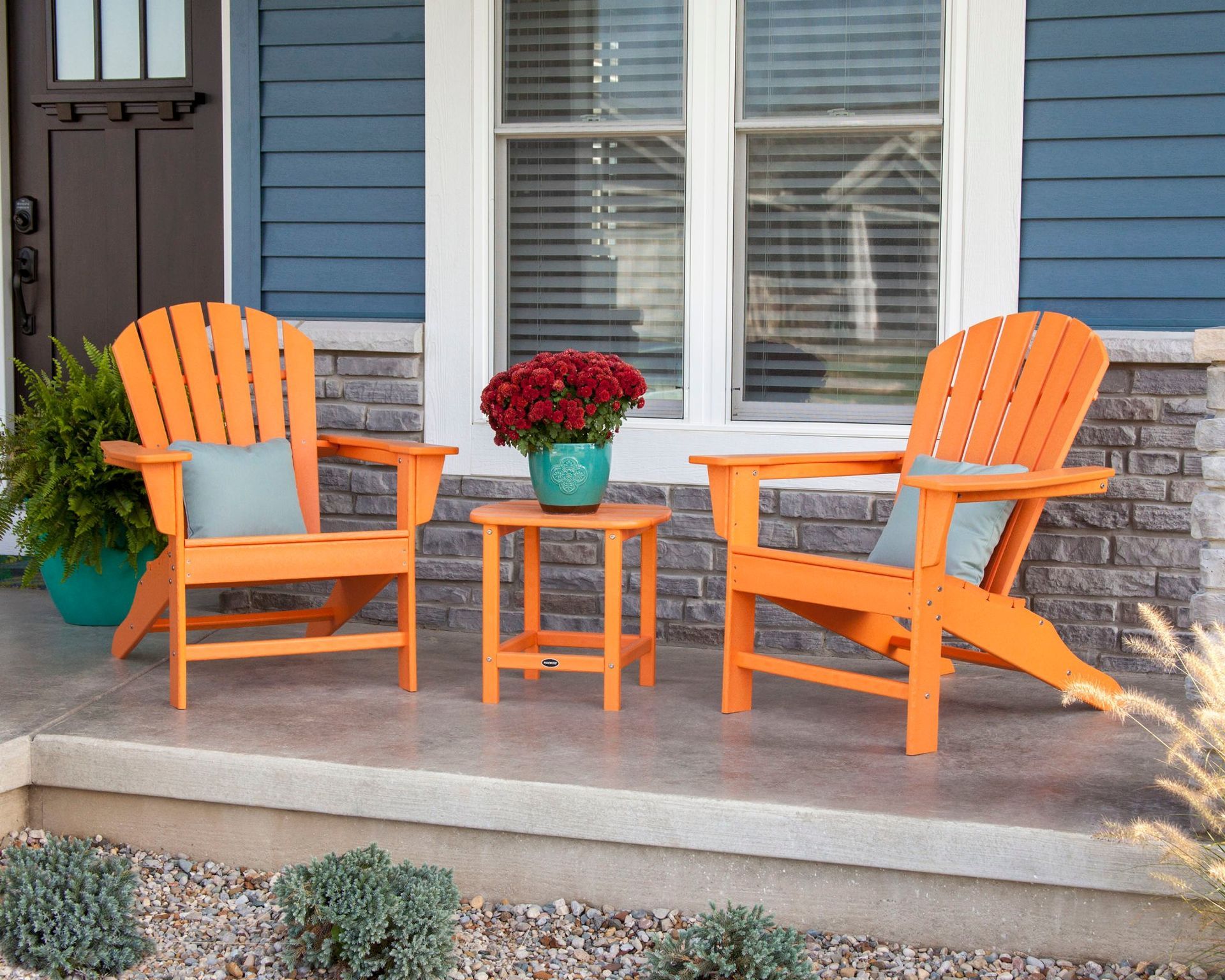Two orange Adirondack chairs and a matching side table sit on a porch with a teal flower pot, against a blue and stone exterior.