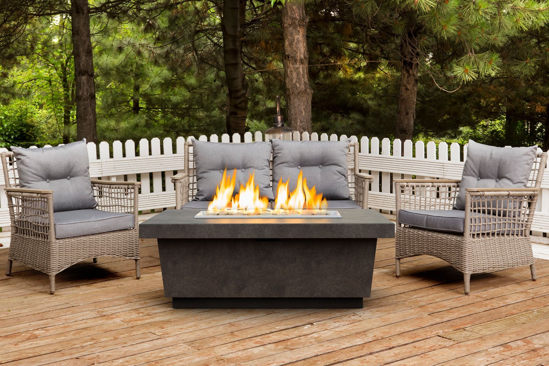 Outdoor seating arrangement with a fire pit table and wicker furniture. Gray cushions, flames, and a wooden deck against a white picket fence and trees.